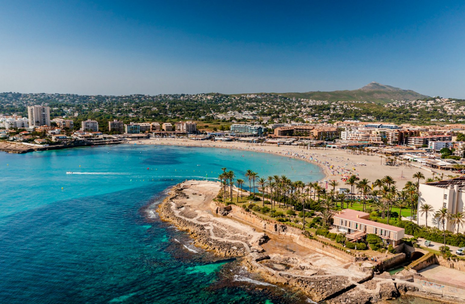 Vista aérea de la Playa El Arenal en Jávea, con su costa rocosa y edificios circundantes bajo un cielo despejado.