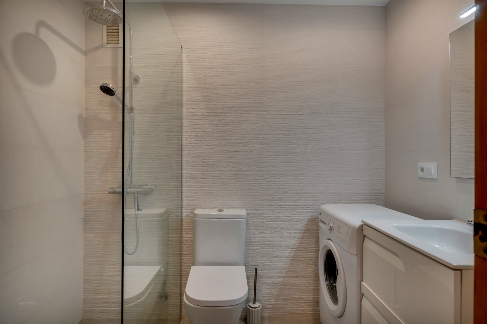 Cuarto de baño con ducha de vidrio, inodoro blanco, lavadora y lavabo con mueble blanco.