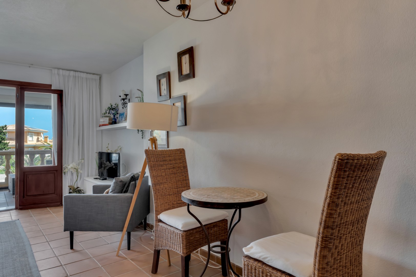 Detalle del salón con mesa de mosaico y sillas de mimbre, iluminado con lámpara de pie.
