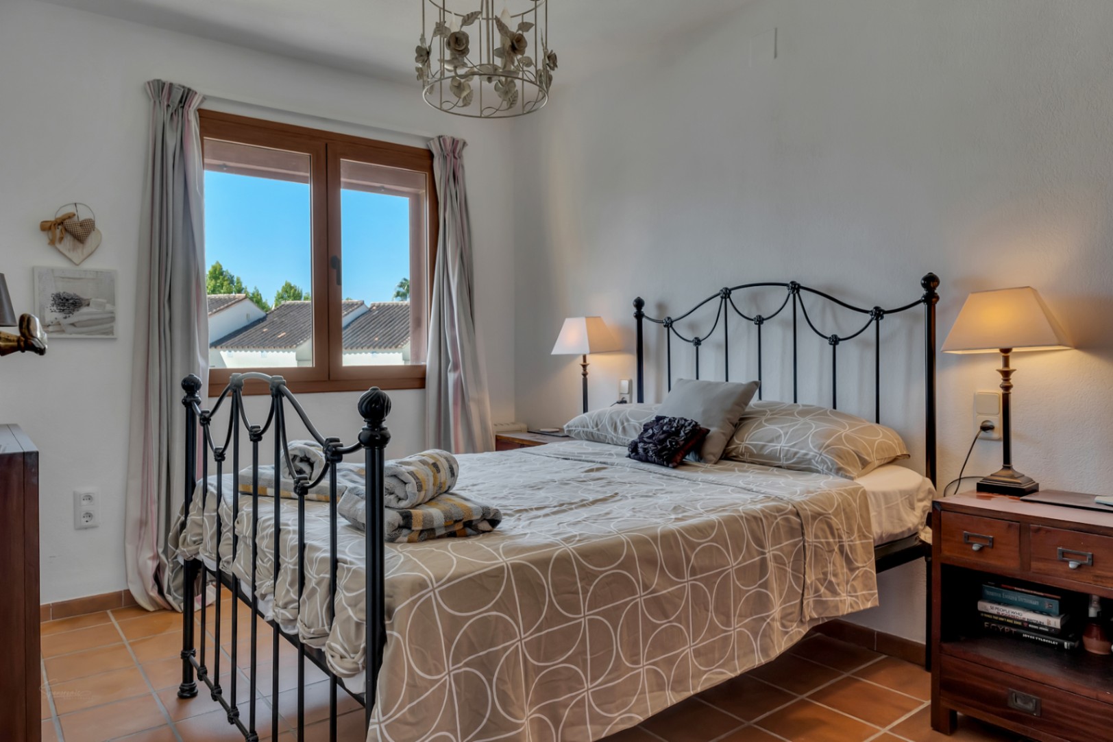 Dormitorio luminoso con cama de hierro forjado, ventana con vistas al exterior y suelo de terracota.