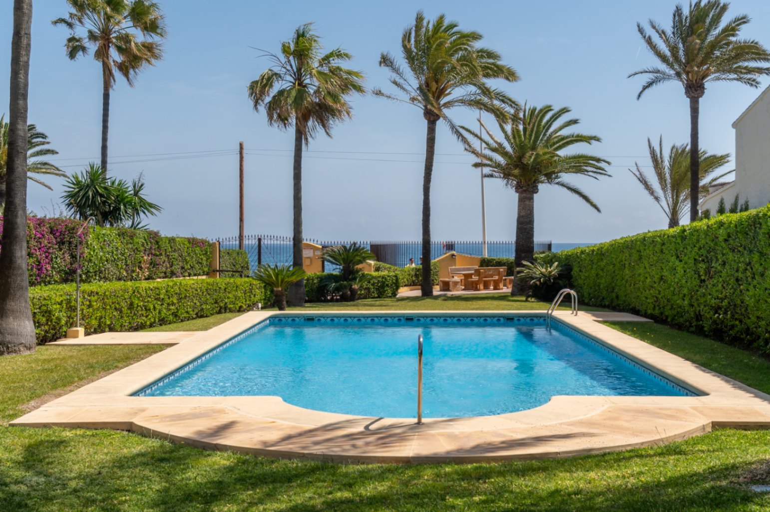 Piscina comunitaria con borde de piedra, rodeada de césped y setos verdes en un jardín mediterráneo.