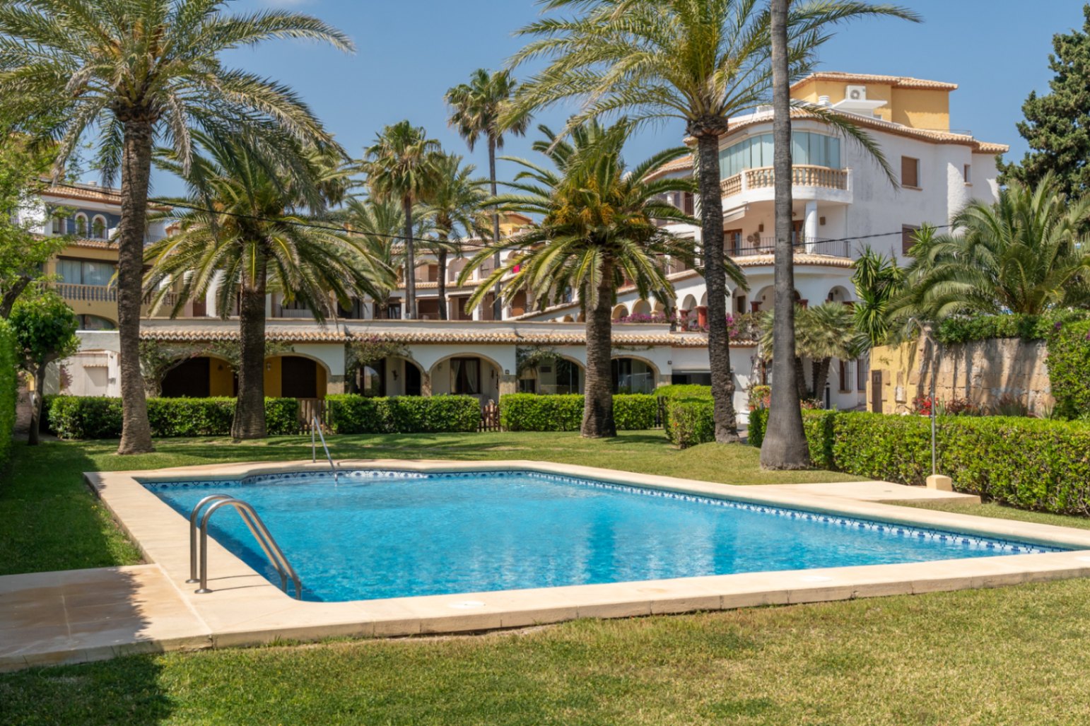 Piscina comunitaria con césped y palmeras en urbanización residencial en Jávea.