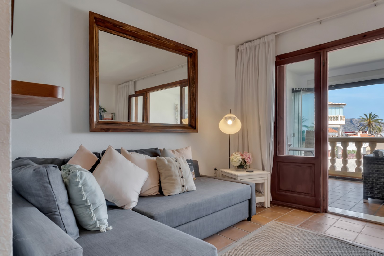 Salón con sofá gris modular, espejo de madera y acceso a terraza con vistas.