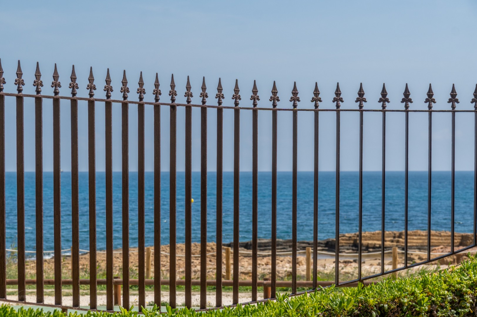 Vista del Mediterráneo a través de una barandilla de hierro ornamental con vegetación en primer plano.
