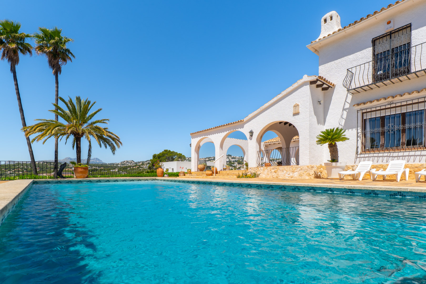 Piscina turquesa con borde de mosaico y villa blanca de estilo mediterráneo con arcos.