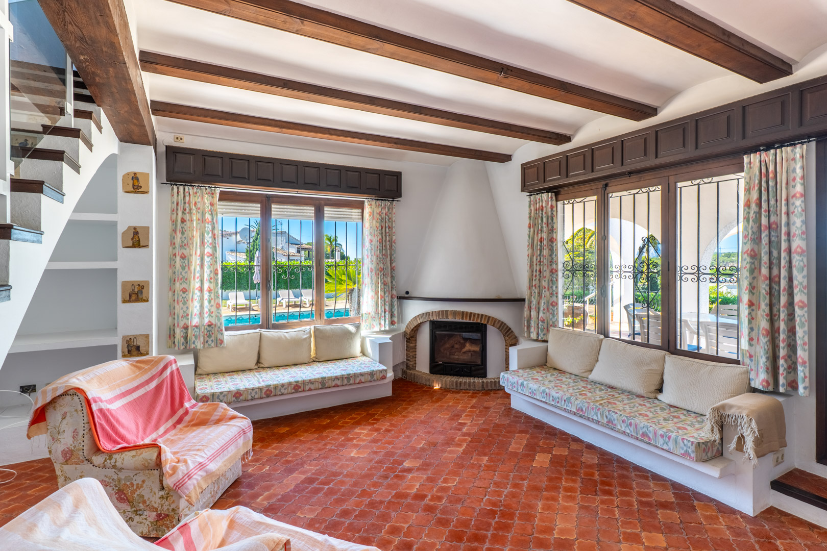 Salón con suelo de terracota, vigas de madera y ventanas con vistas a la piscina.