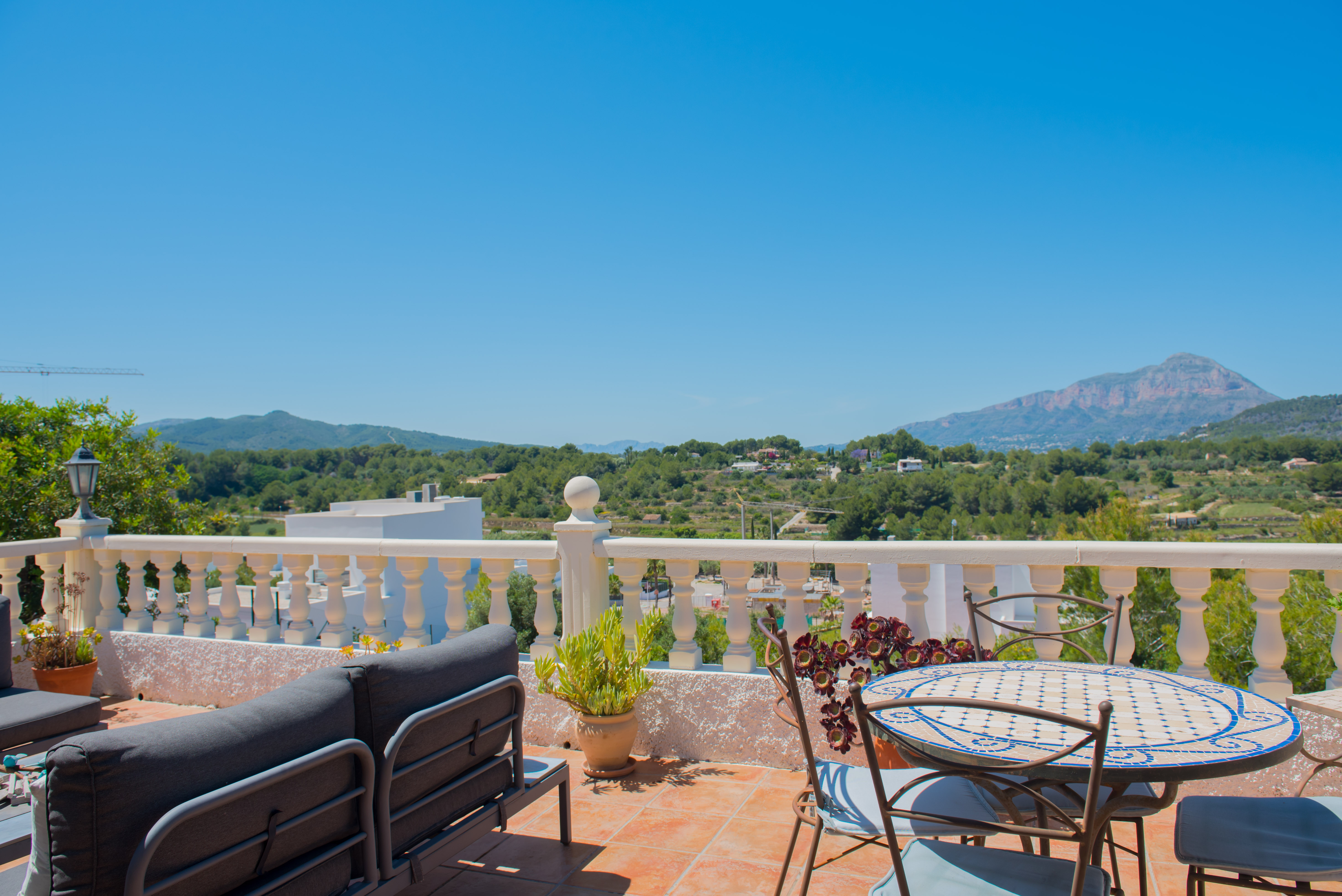 Terraza con baldosas y vistas a la montaña, amueblada con un sofá gris y una mesa de mosaico.