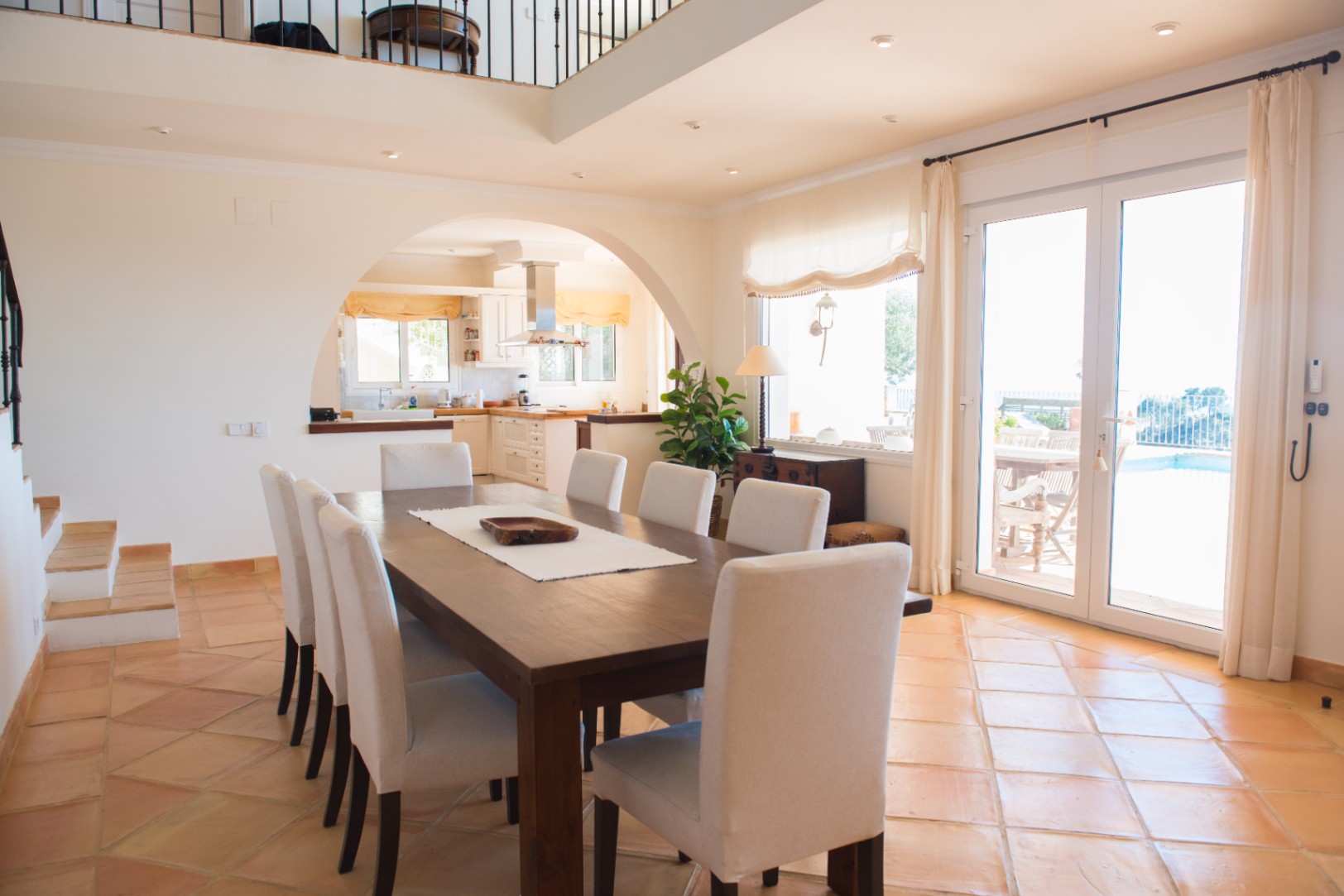 Comedor espacioso con mesa de madera maciza, suelos de terracota y un arco hacia la cocina, con acceso directo a la terraza exterior a través de ventanales.