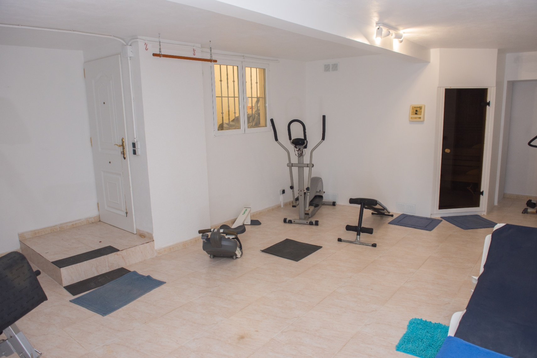 Gimnasio privado con máquinas de cardio, banco de pesas y acceso directo a una sauna. La estancia cuenta con suelos cerámicos y paredes blancas.