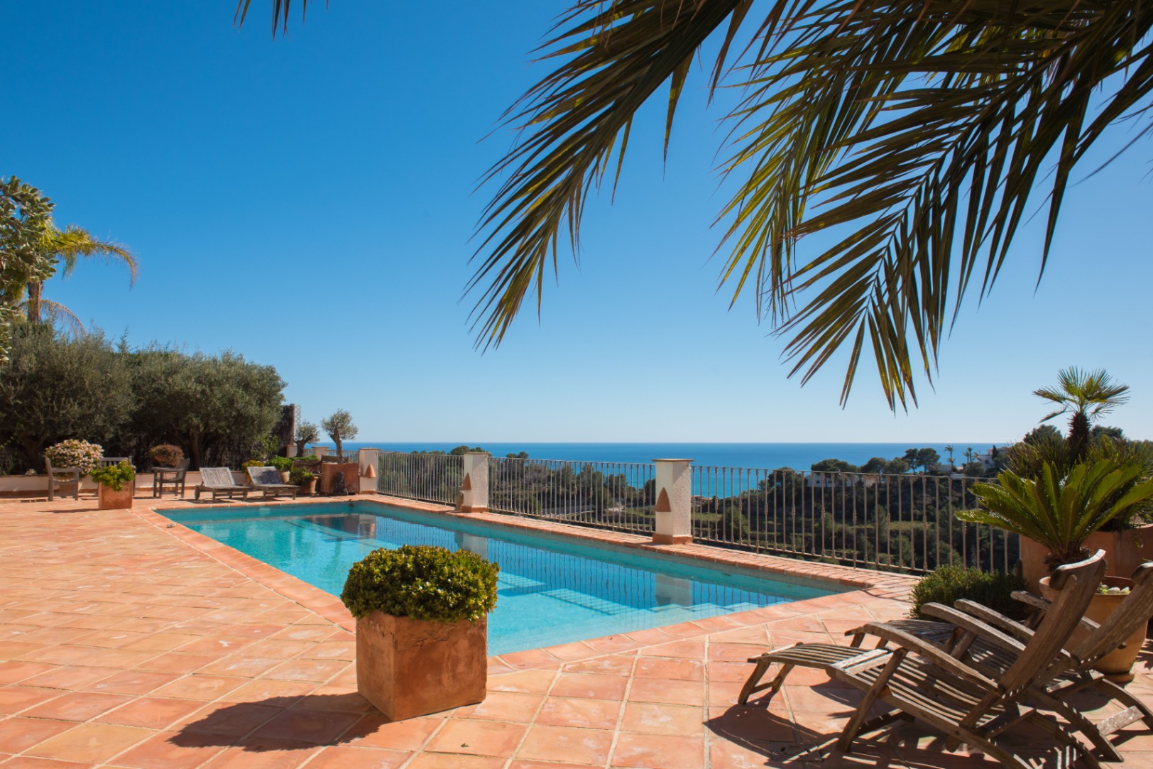 Terraza de baldosas de terracota con piscina rectangular, tumbonas de madera y vistas panorámicas al mar Mediterráneo bajo un cielo despejado.