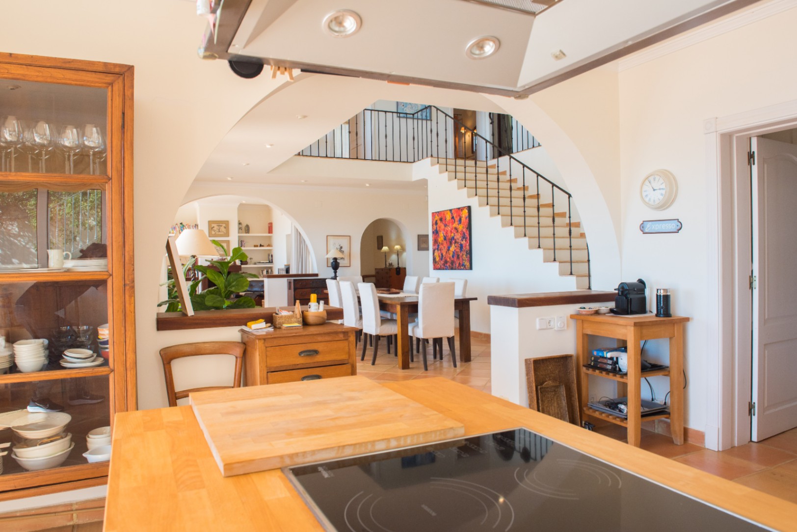 Vista desde la isla de cocina con encimera de madera hacia el comedor y la escalera principal, destacando los arcos arquitectónicos y suelos cerámicos.