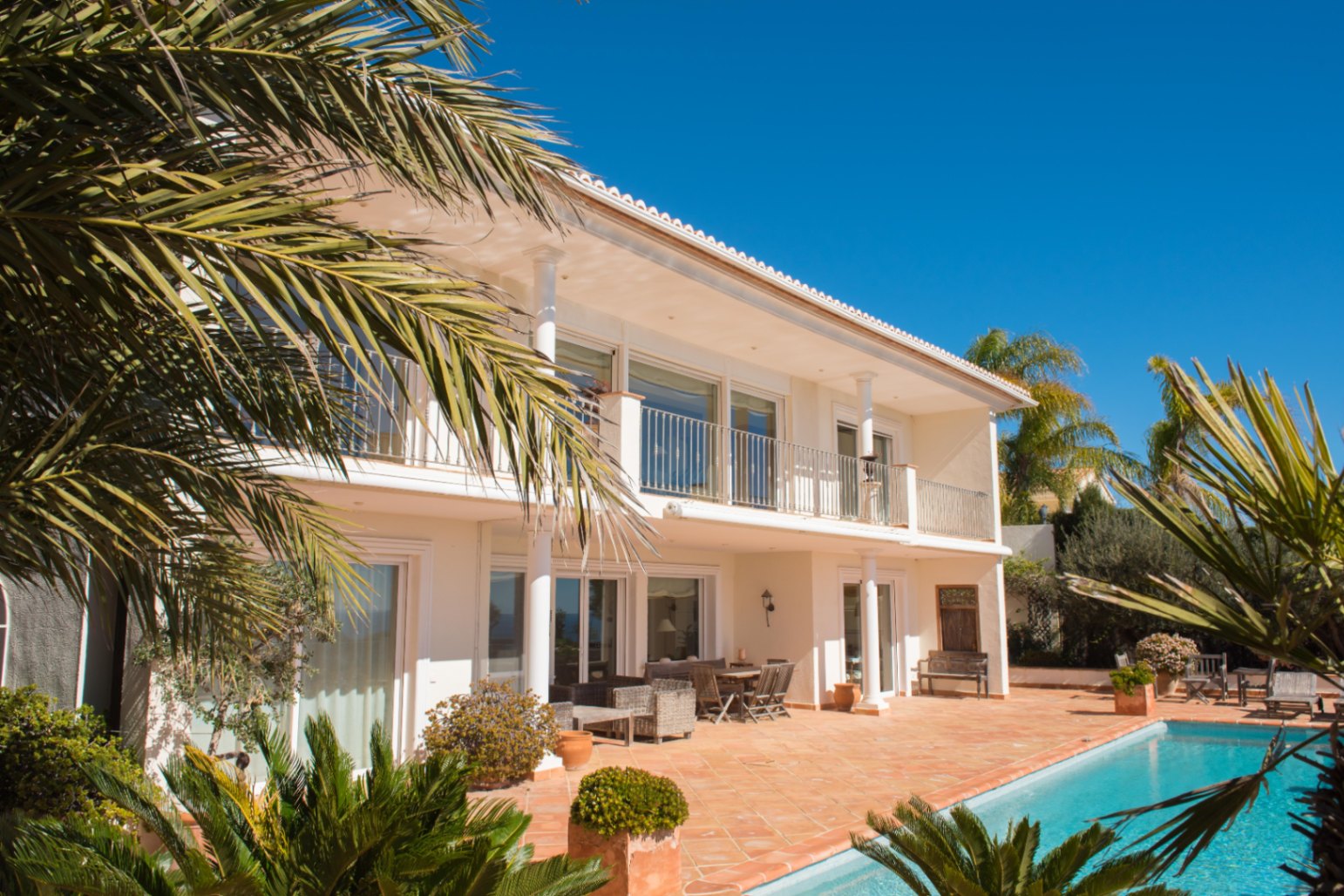 Vista exterior de la villa de dos plantas con terraza de terracota, piscina privada y balcón superior con columnas blancas y barandillas metálicas.