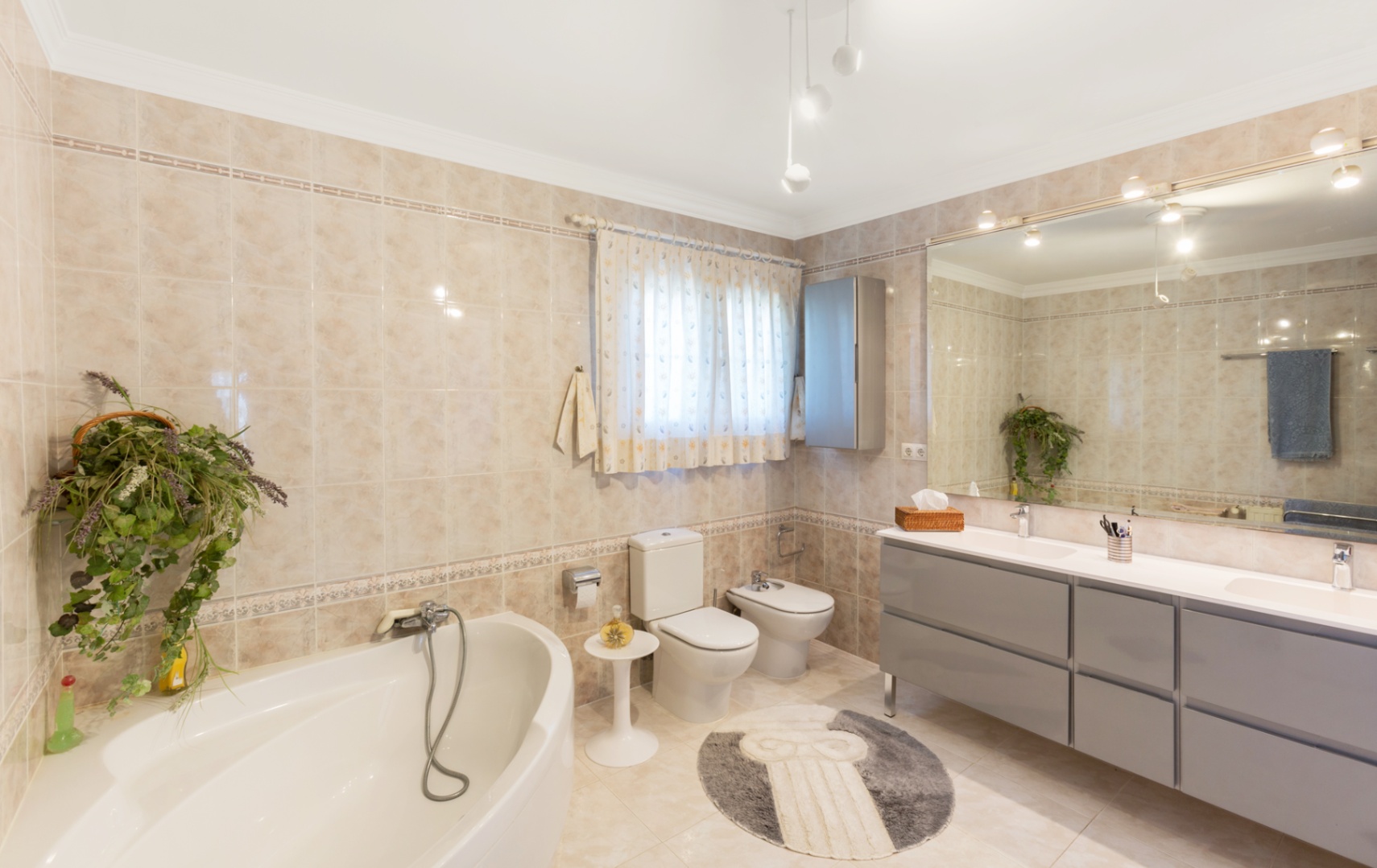 Baño espacioso equipado con bañera de rincón, mueble de doble lavabo en gris y un gran espejo. Revestimiento de azulejos beige con cenefas decorativas.