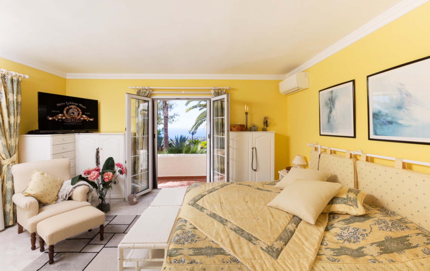 Dormitorio con paredes amarillas, mobiliario blanco y zona de estar. Las puertas dobles de cristal conectan con una terraza con vistas al mar y vegetación mediterránea.