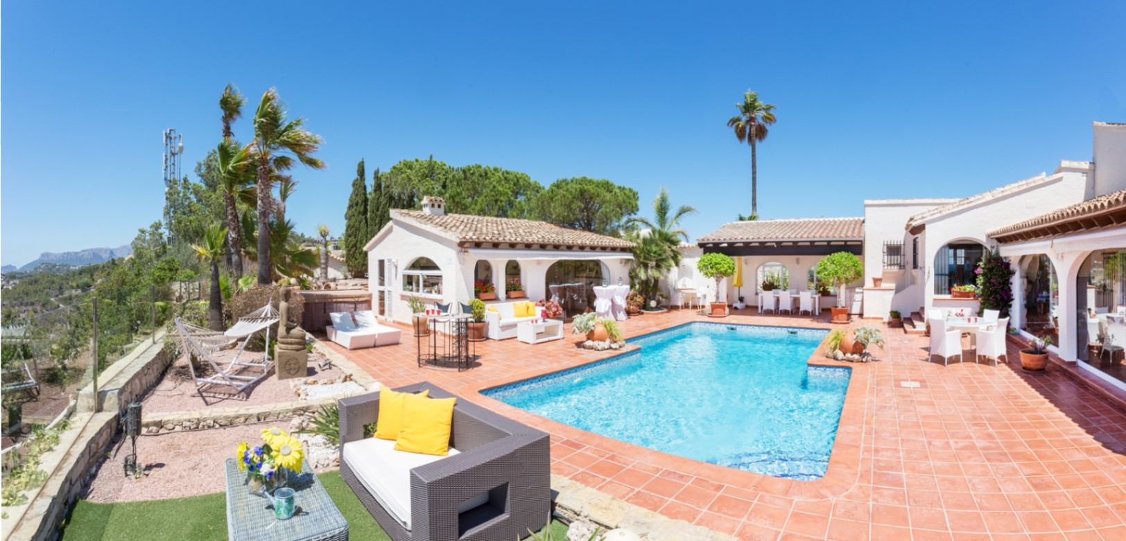 Terraza con piscina rectangular, suelo de terracota y varias zonas de estar. La arquitectura mediterránea destaca por sus paredes blancas y ventanas de arco.