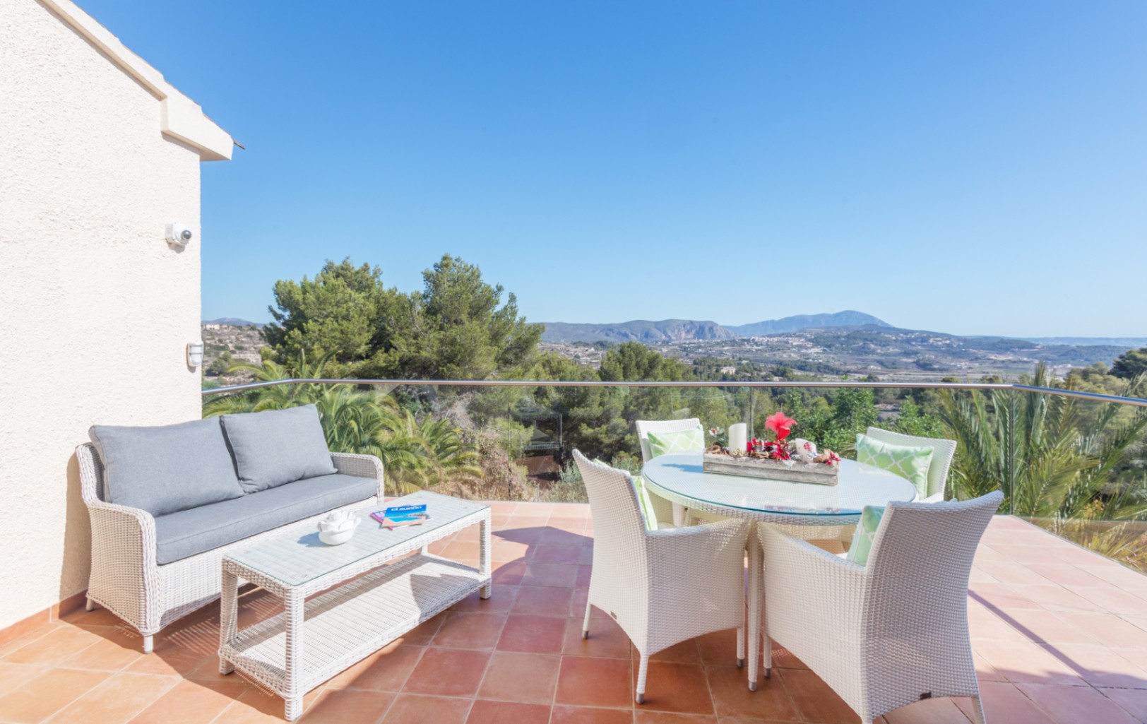 Terraza con suelo de terracota, mobiliario de ratán blanco y barandilla de cristal con vistas panorámicas a las montañas y el entorno natural de Benitachell.