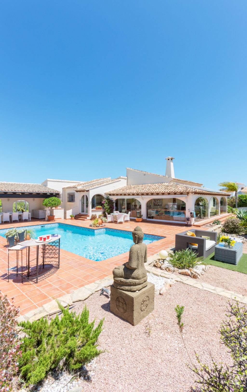 Terraza exterior con piscina de azulejos azules, suelos de terracota, una estatua de Buda de piedra y villa mediterránea con porche acristalado.