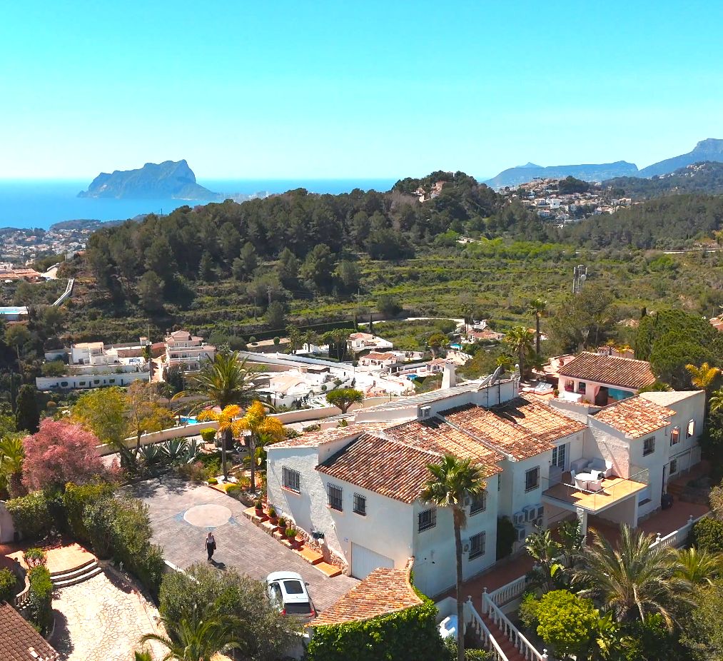 Vista aérea de la villa mediterránea con tejados de terracota, jardines cuidados y vistas panorámicas al mar y al Peñón de Ifach en el horizonte.