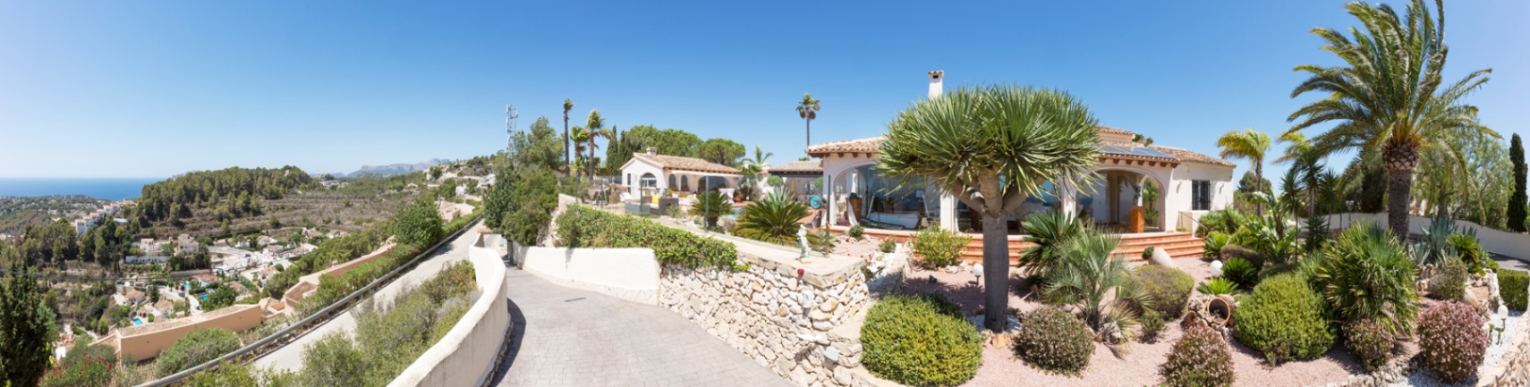 Vista panorámica del exterior de la villa con entrada pavimentada, muros de piedra seca y un jardín mediterráneo con palmeras y vistas despejadas a la costa.