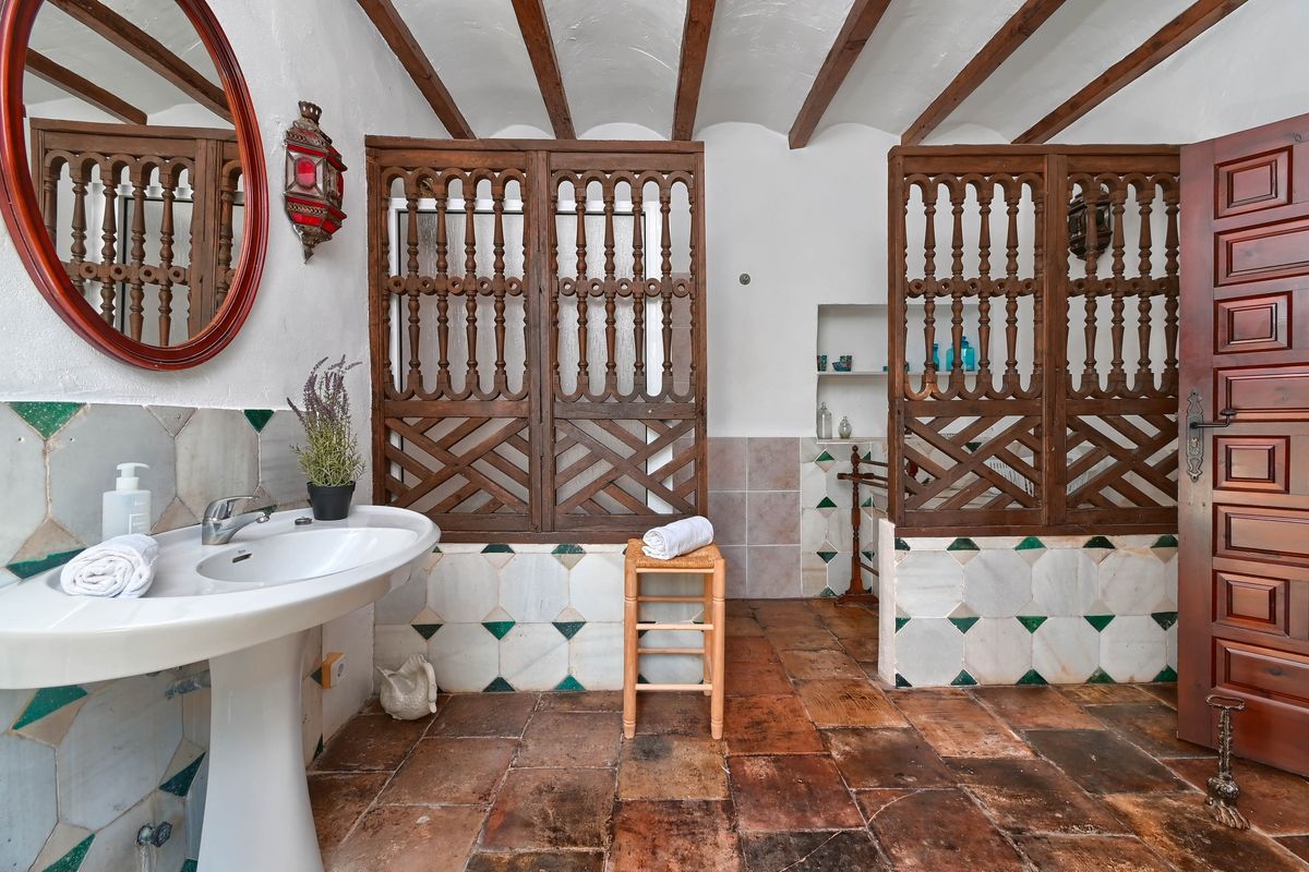 Baño rústico con vigas de madera vistas, suelos de barro cocido, lavabo de pedestal y celosías de madera tallada de estilo tradicional.