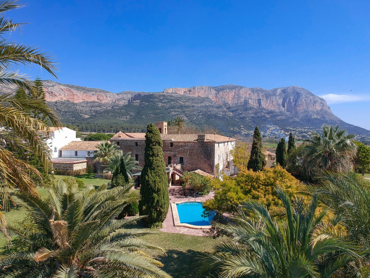 Finca histórica de piedra con piscina rodeada de jardines mediterráneos y vistas panorámicas al macizo del Montgó bajo un cielo despejado.