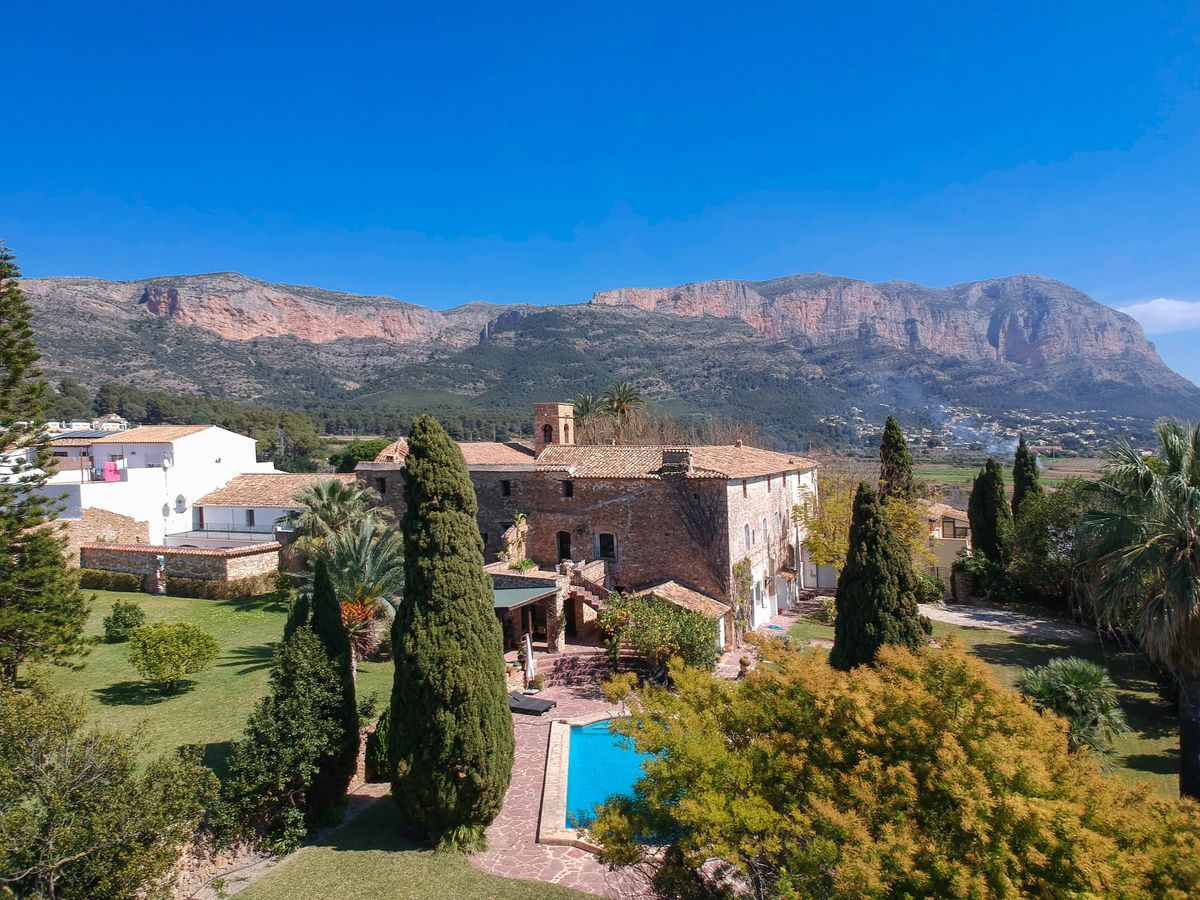 Vista elevada de la finca histórica de piedra con piscina, jardines cuidados con cipreses y el imponente macizo del Montgó al fondo.