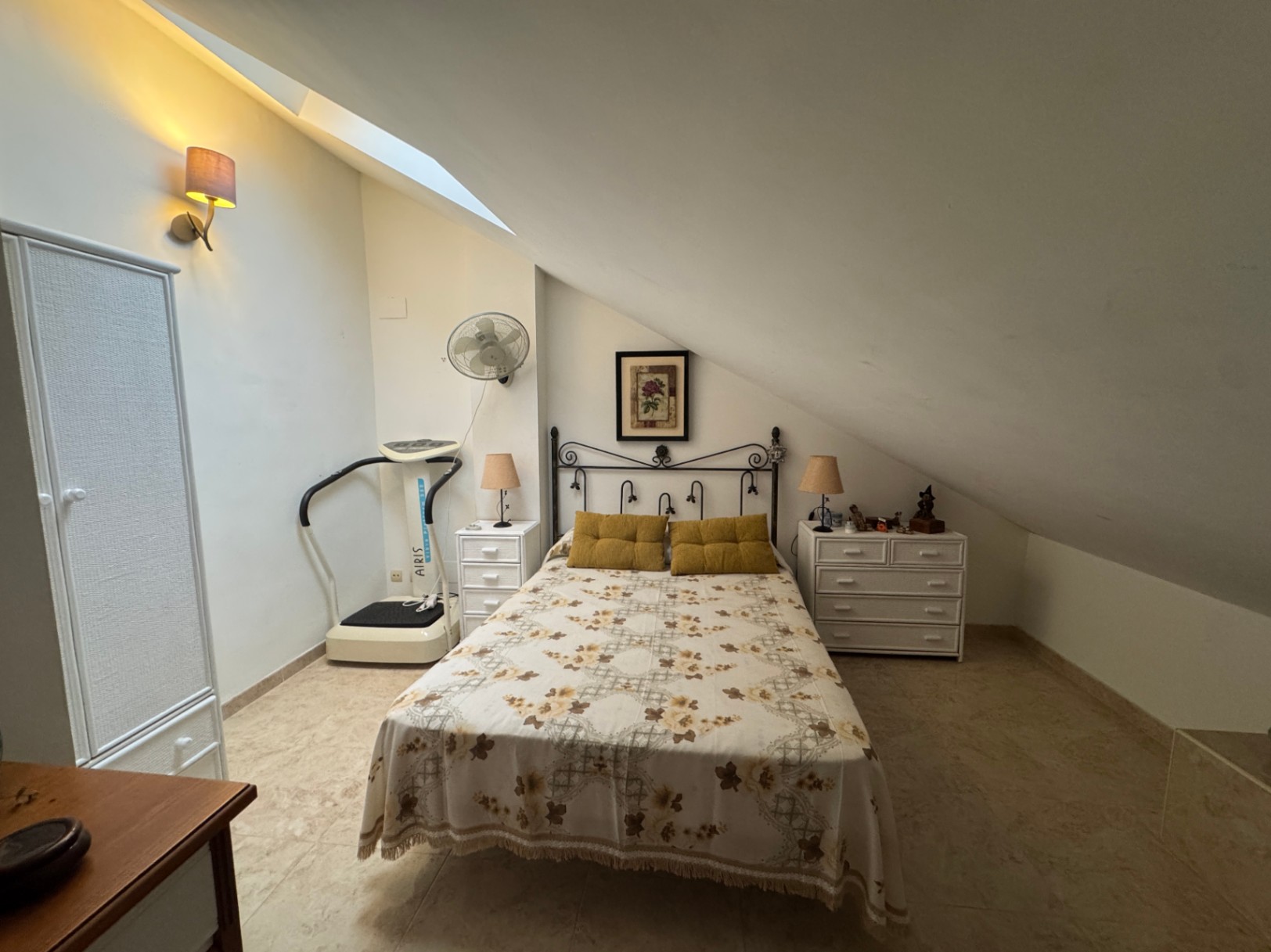 Dormitorio abuhardillado con cama doble, máquina de ejercicios y armarios blancos.
