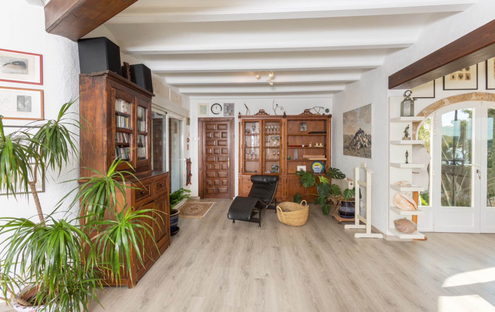 Amplia zona de estar con techos de vigas blancas, muebles de madera rústica y suelos laminados claros. La luz natural entra por puertas acristaladas con arcos de piedra.