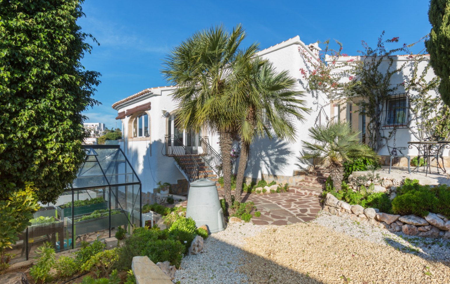 Exterior de una villa mediterránea blanca con senderos de piedra, palmeras maduras y un invernadero de cristal integrado en el jardín paisajístico.