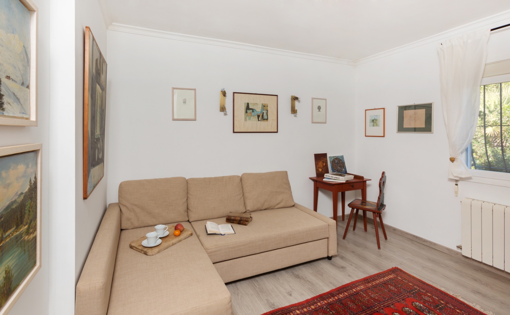 Zona de estar luminosa con sofá chaise longue beige, escritorio de madera clásico y paredes decoradas con obras de arte sobre suelo laminado gris.