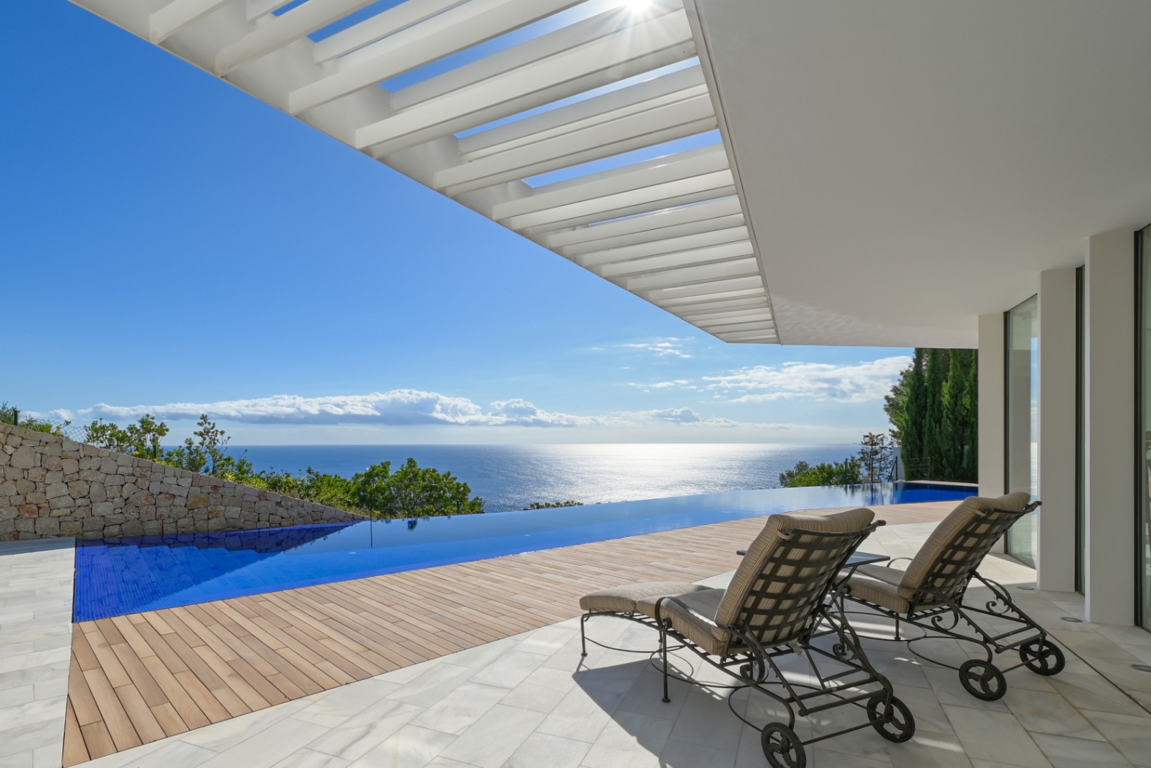 Terraza con piscina infinita y vistas panorámicas al mar, suelo de mármol y madera bajo una pérgola blanca con tumbonas de hierro forjado.