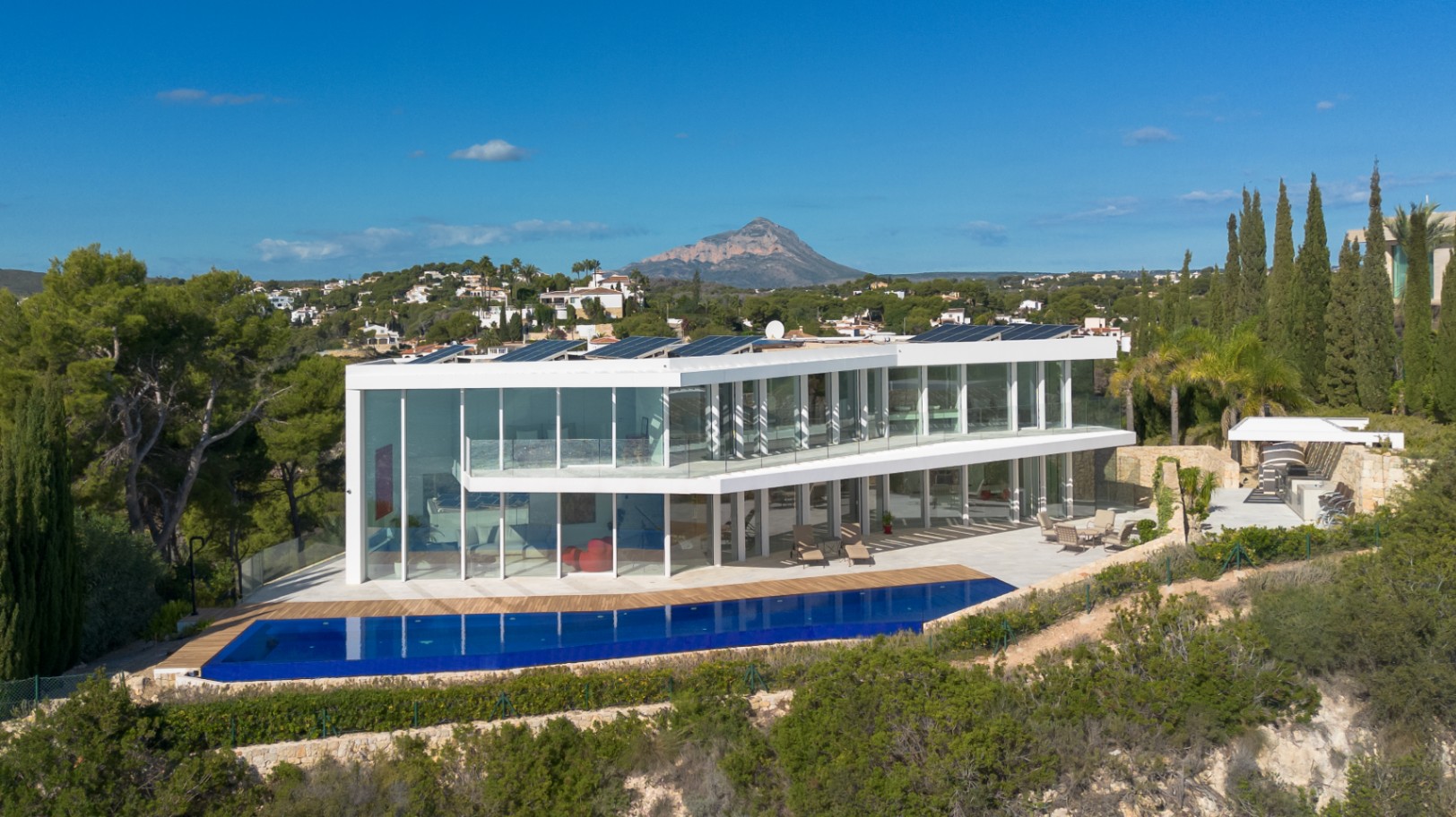 Villa moderna de dos niveles con fachadas acristaladas, piscina infinita, paneles solares y vistas despejadas al macizo del Montgó.