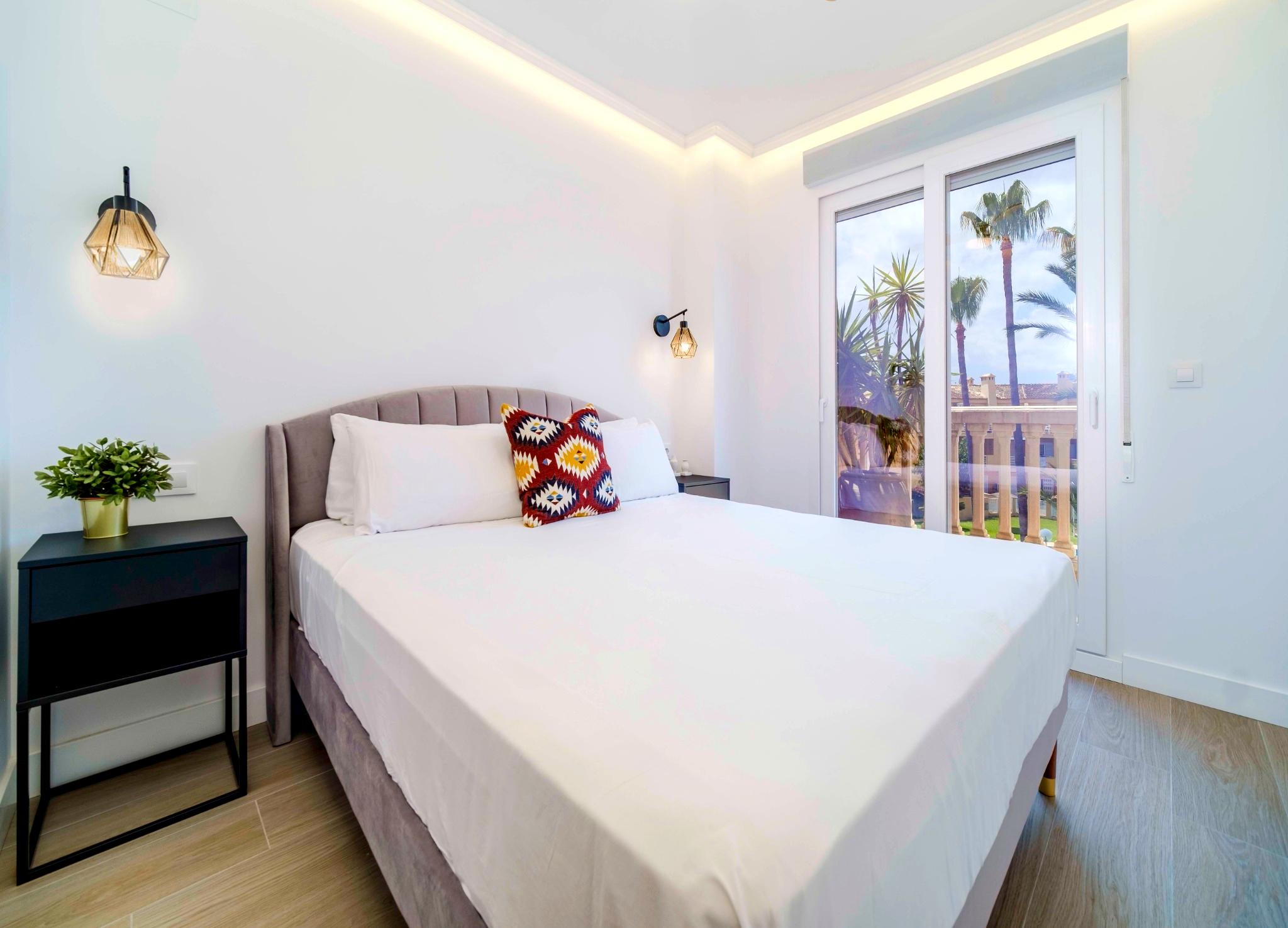 Dormitorio moderno con cabecero de terciopelo, apliques geométricos e iluminación LED indirecta. Grandes ventanales dan acceso a un balcón con vistas a las palmeras.