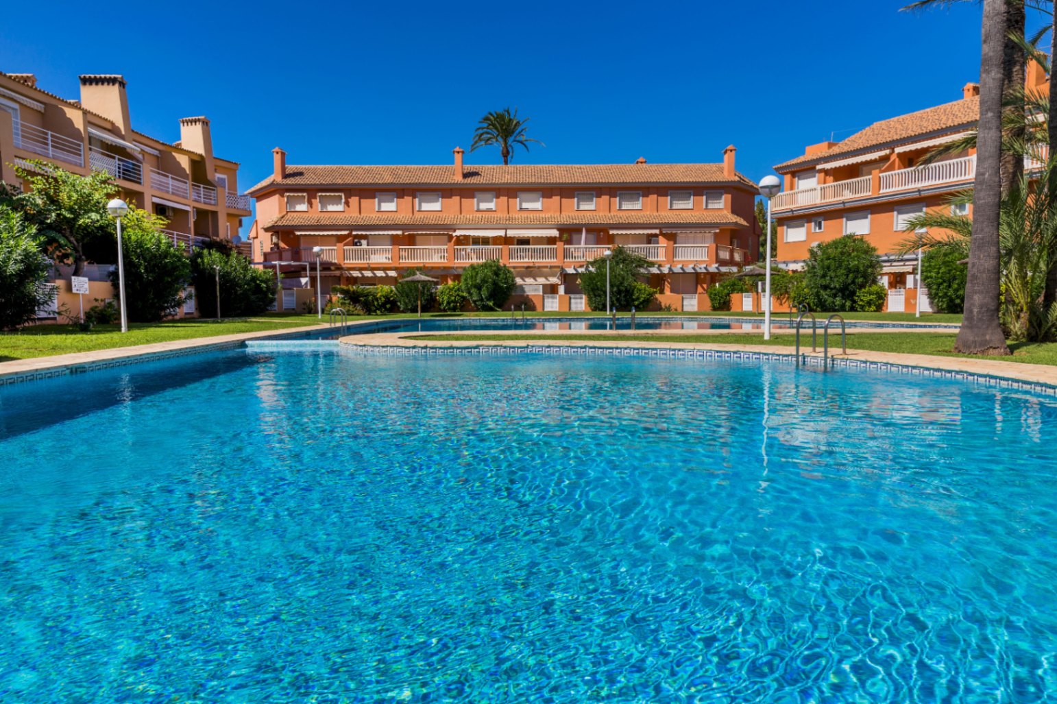Piscina comunitaria de gresite azul rodeada de zonas verdes y edificios residenciales de color terracota con amplios balcones y palmeras.