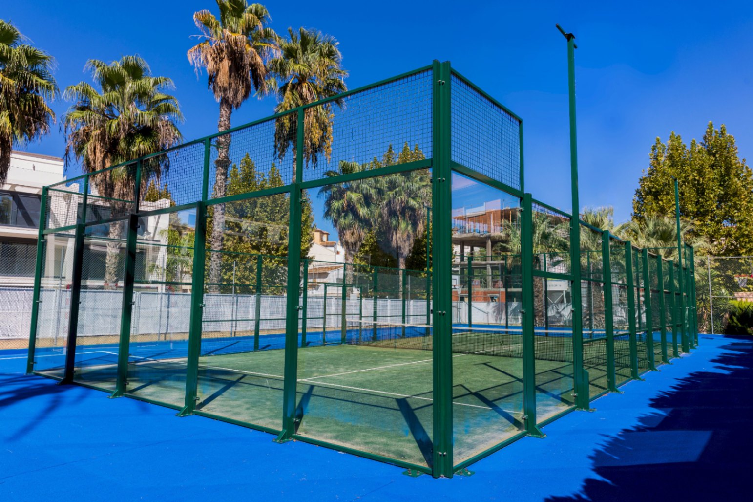 Pista de pádel exterior con césped artificial verde, cerramientos de vidrio templado y estructura metálica, rodeada de palmeras y superficie azul.