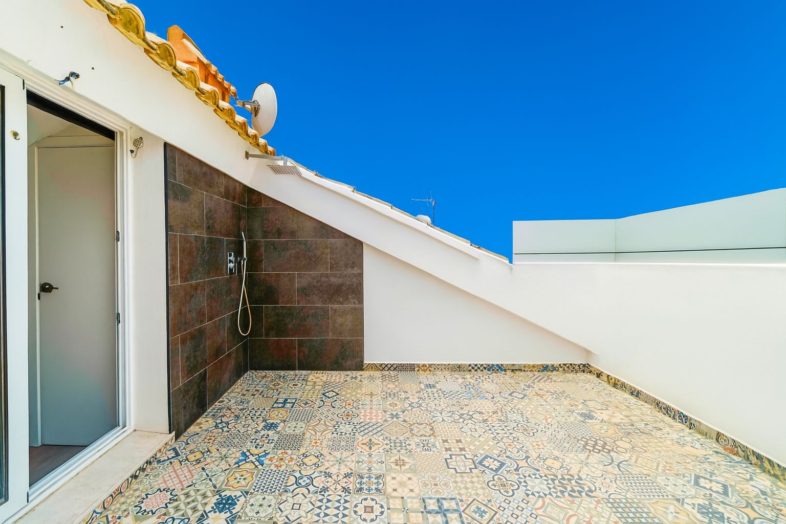 Terraza en la azotea con suelo de baldosas hidráulicas decorativas, ducha exterior sobre pared de gres oscuro y barandilla de cristal.