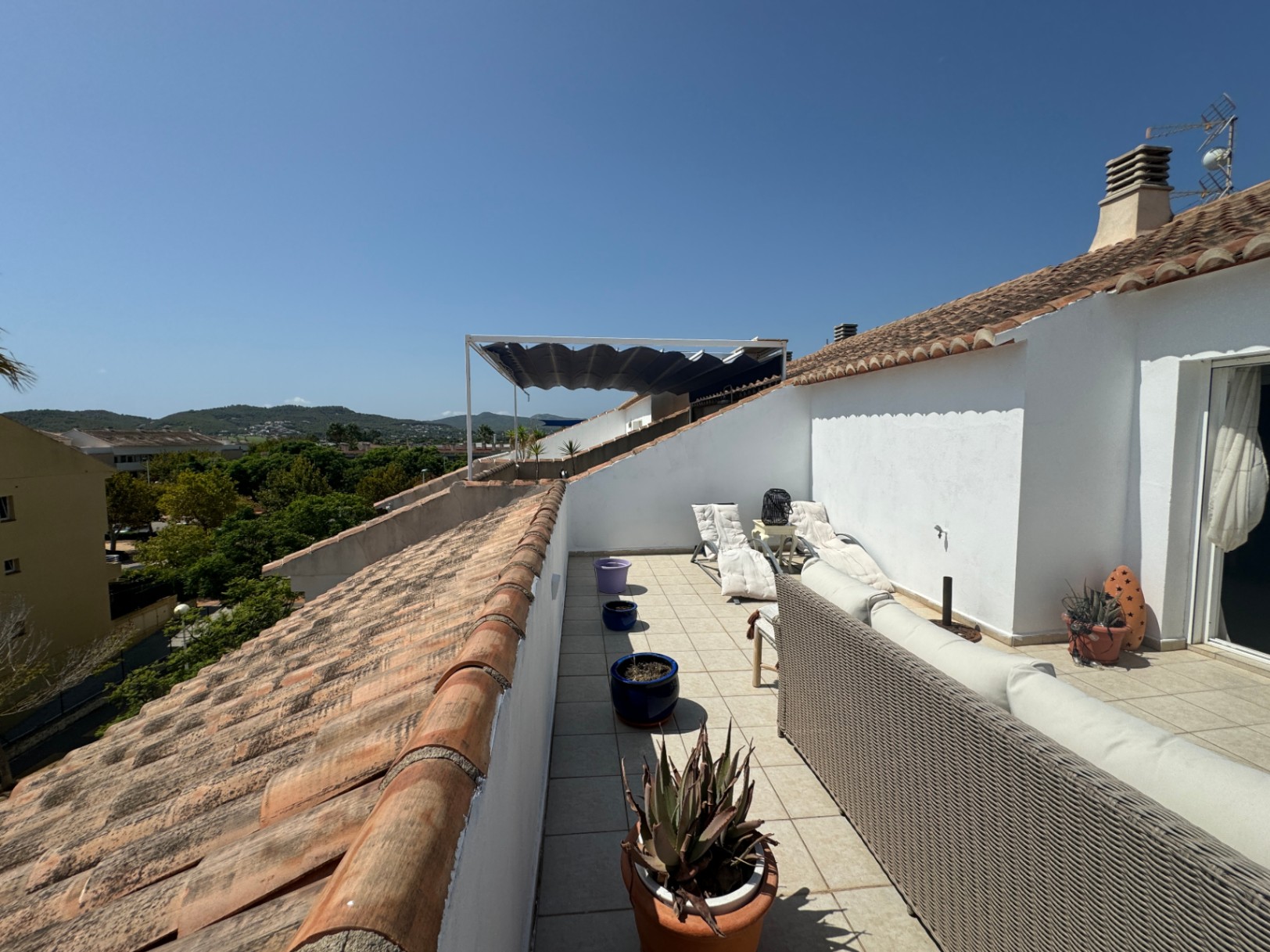 Terraza alargada en la azotea con sofá de mimbre gris, tumbonas blancas y suelos cerámicos, con vistas despejadas a las colinas y tejados de teja tradicional.