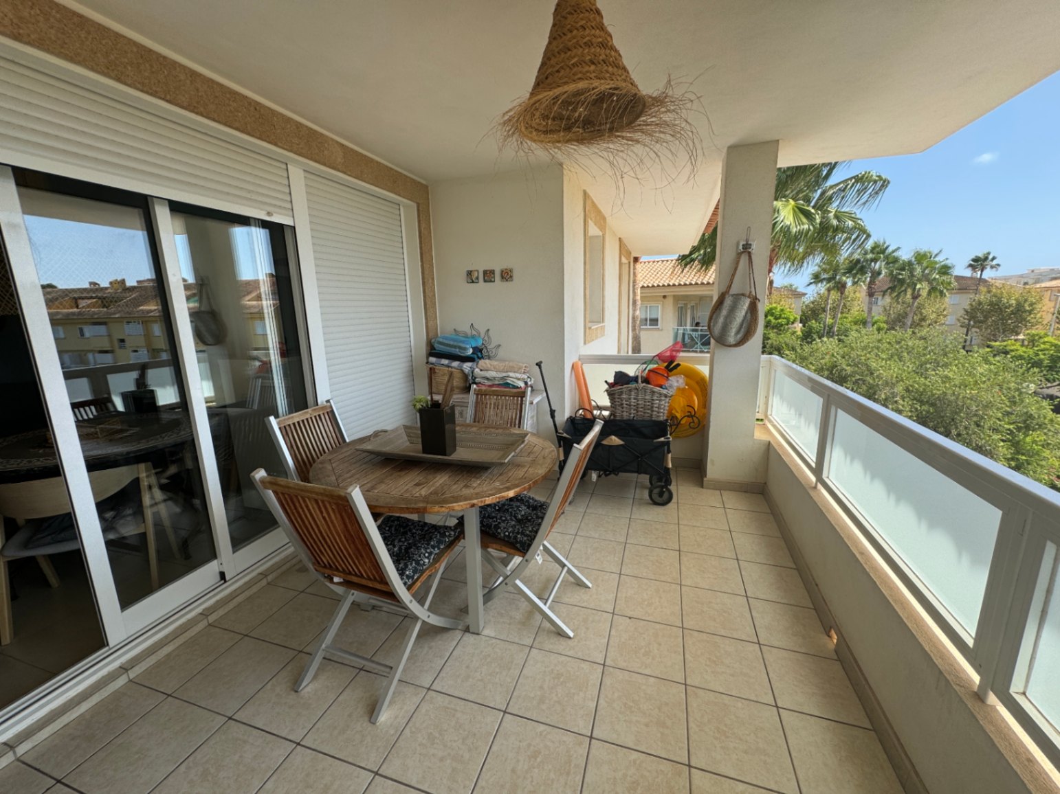 Terraza cubierta con conjunto de comedor de madera, suelo cerámico y barandilla de cristal con vistas a palmeras y zonas residenciales.
