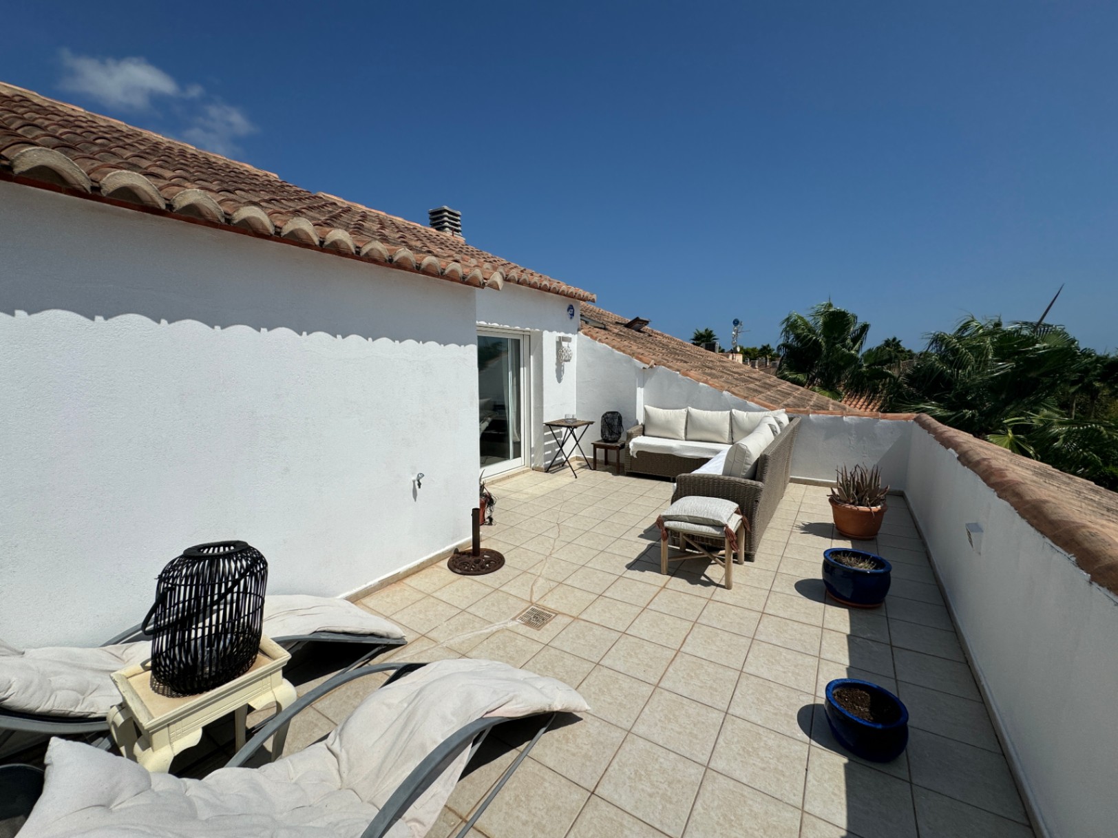 Terraza en azotea con paredes de estuco blanco, tejado de teja, mobiliario de mimbre y suelo cerámico con vistas a palmeras.