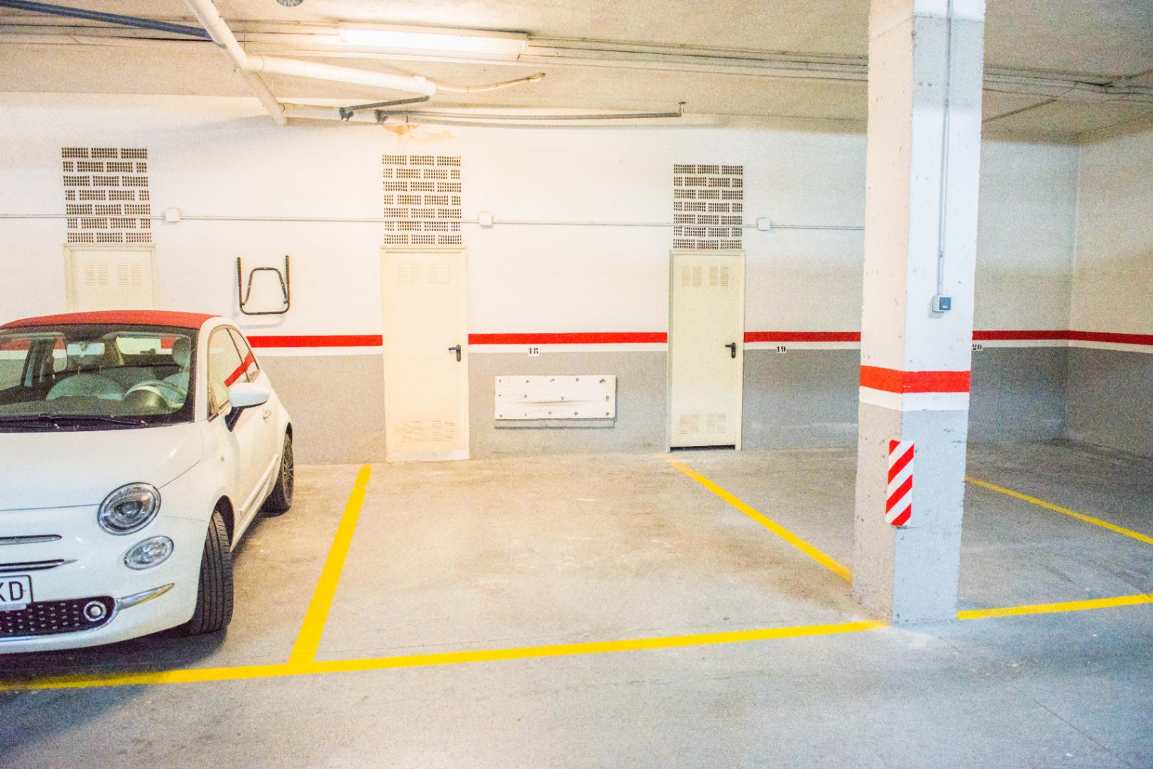 Plaza de garaje subterránea con numeración, líneas amarillas de delimitación y paredes en tonos blanco y gris con una franja roja decorativa.