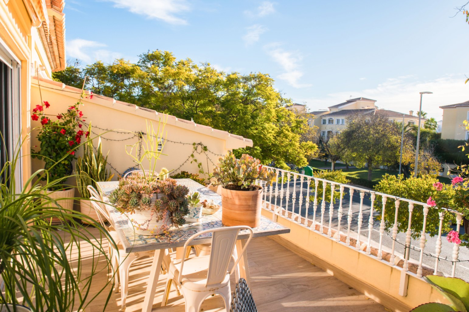 Terraza con mesa de comedor de mosaico, sillas metálicas blancas y plantas suculentas con vistas a una zona residencial y árboles frondosos.