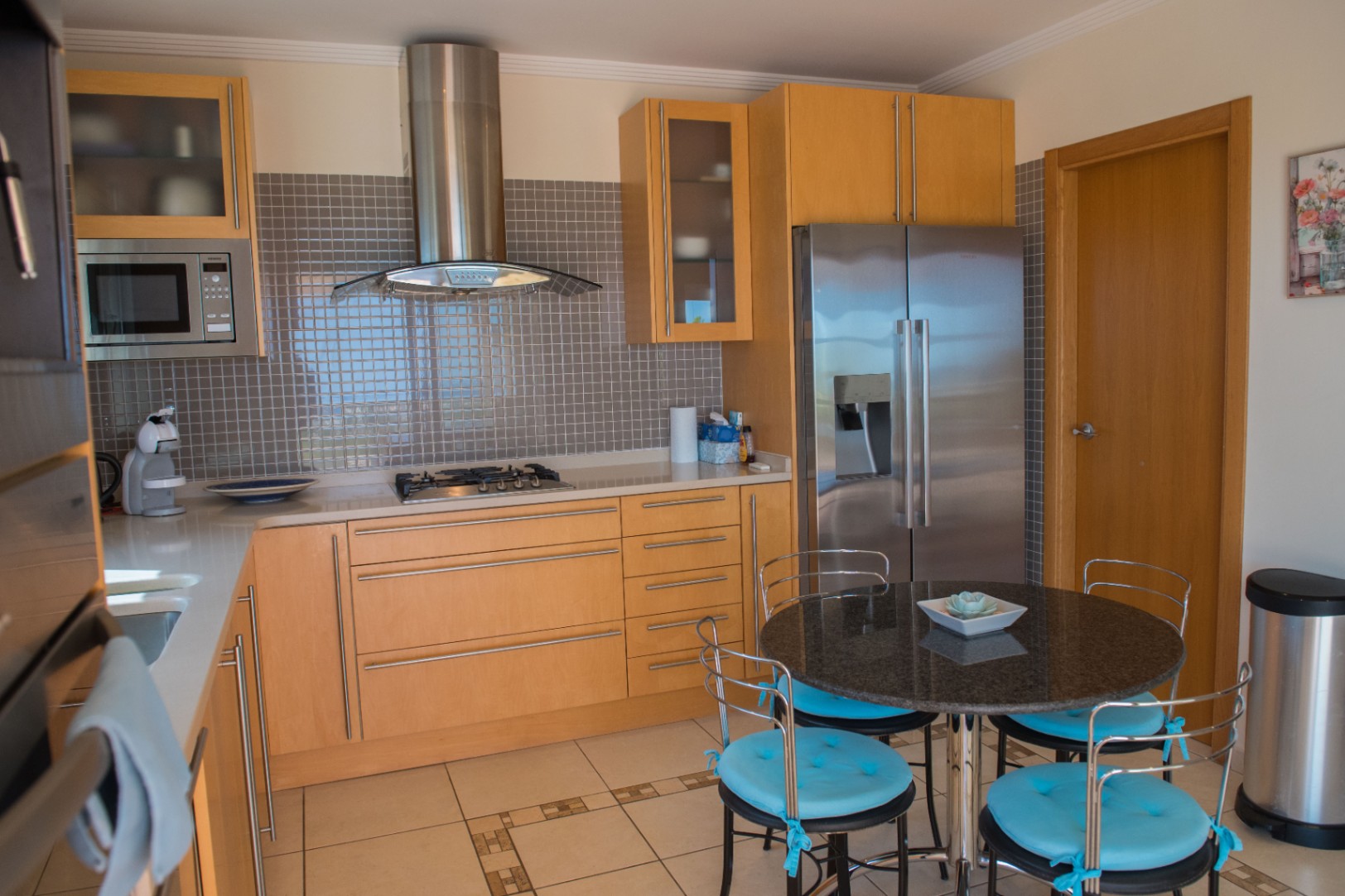 Cocina moderna con armarios de madera clara, salpicadero de mosaico gris y electrodomésticos de acero inoxidable. Incluye una mesa redonda de granito con sillas de cojines azules.