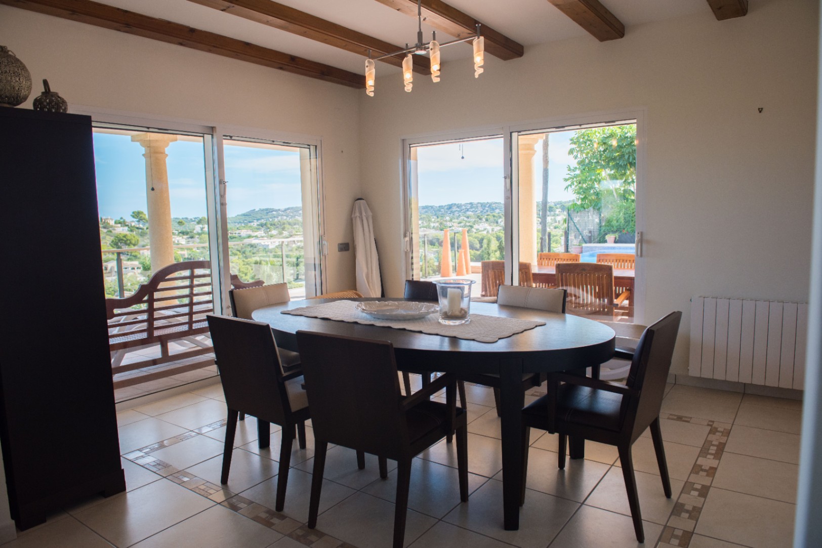 Comedor con mesa ovalada de madera oscura y vigas a la vista. Grandes ventanales correderos ofrecen vistas panorámicas al valle y acceso directo a la terraza exterior.