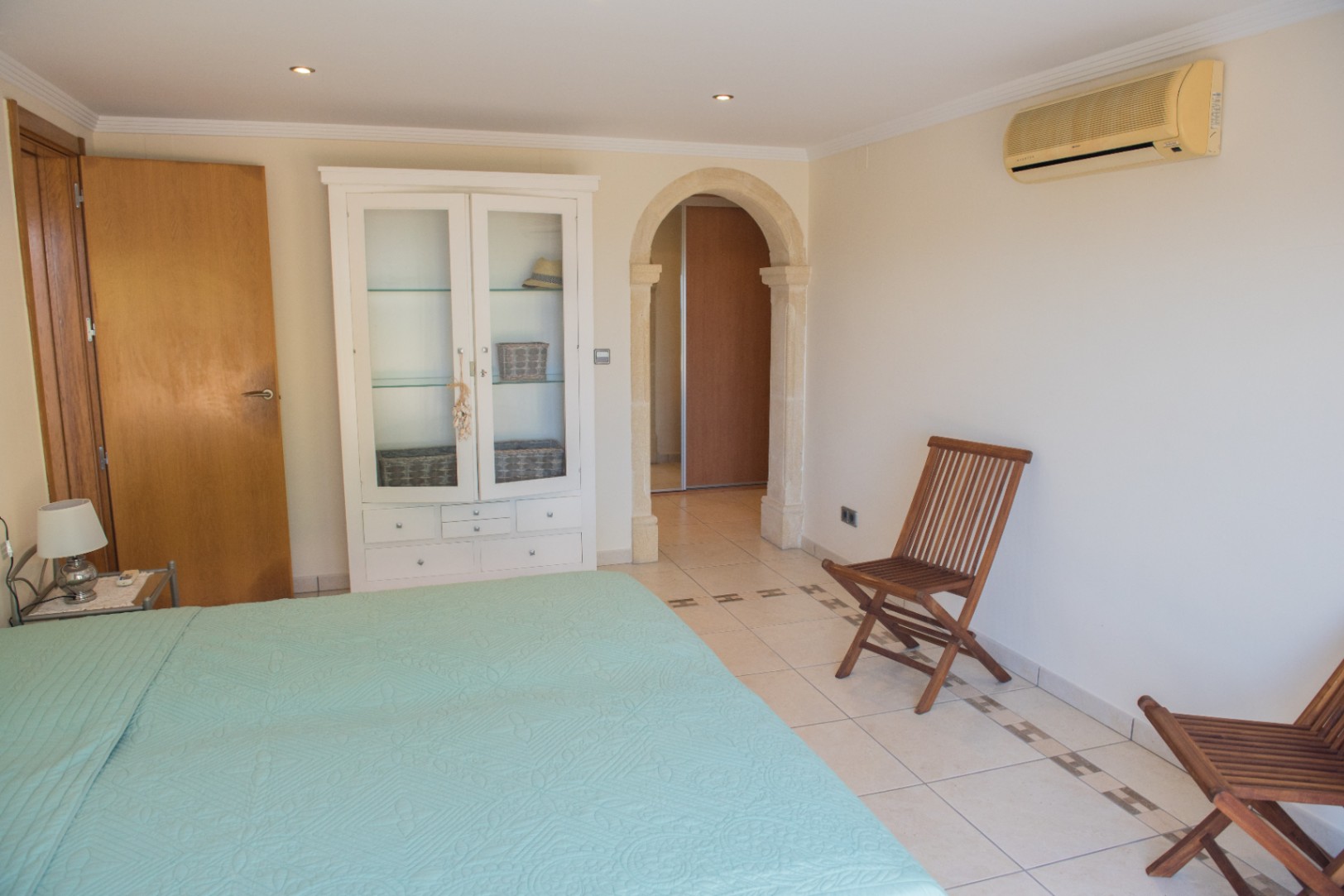 Dormitorio con arco de piedra natural, suelos cerámicos con cenefa decorativa, armario vitrina blanco y sillas plegables de madera. Incluye aire acondicionado y luces empotradas.