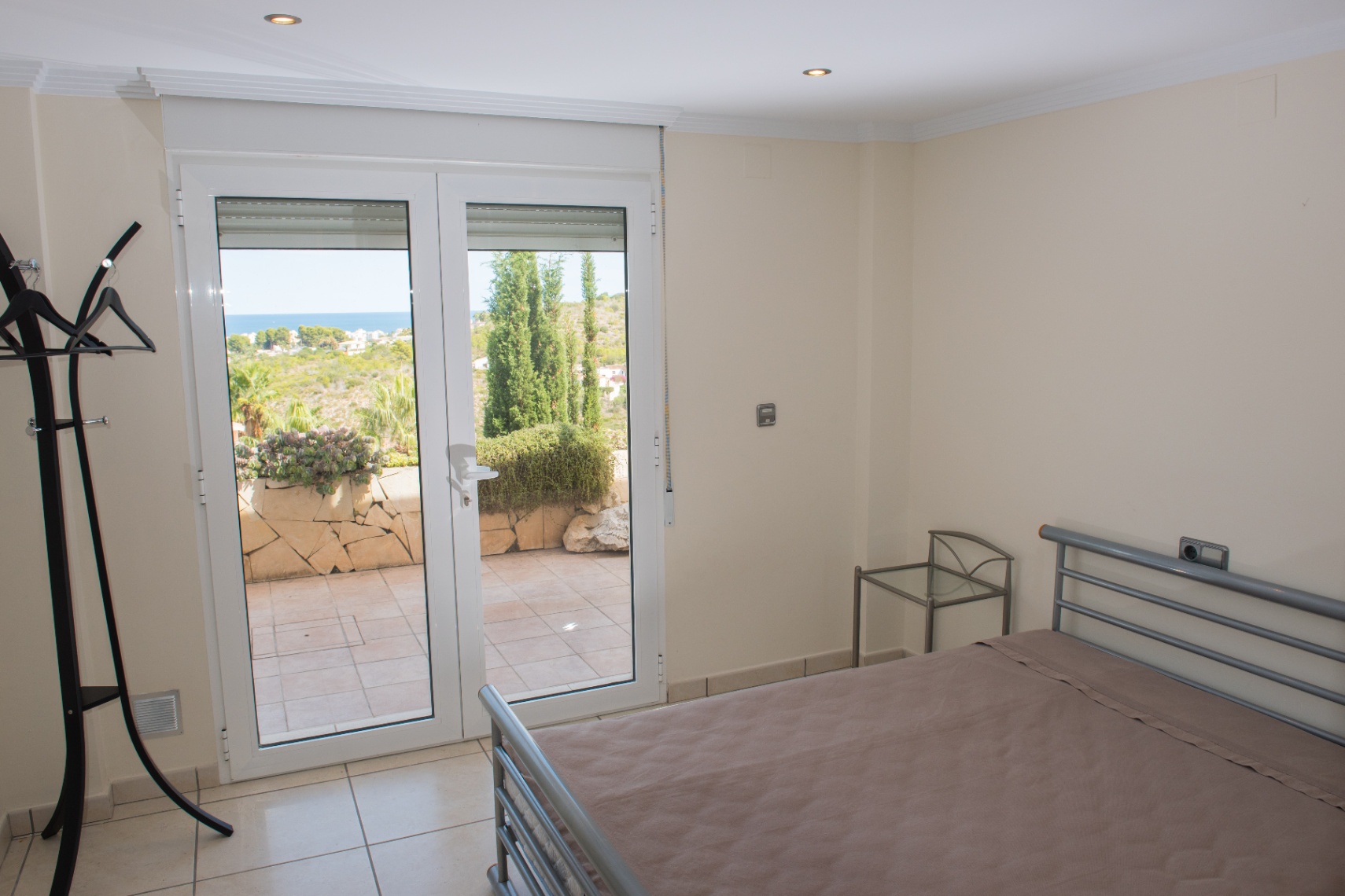 Dormitorio con cama de metal gris y mesa de cristal, con puertas dobles que dan a una terraza solada con vistas despejadas a la costa.