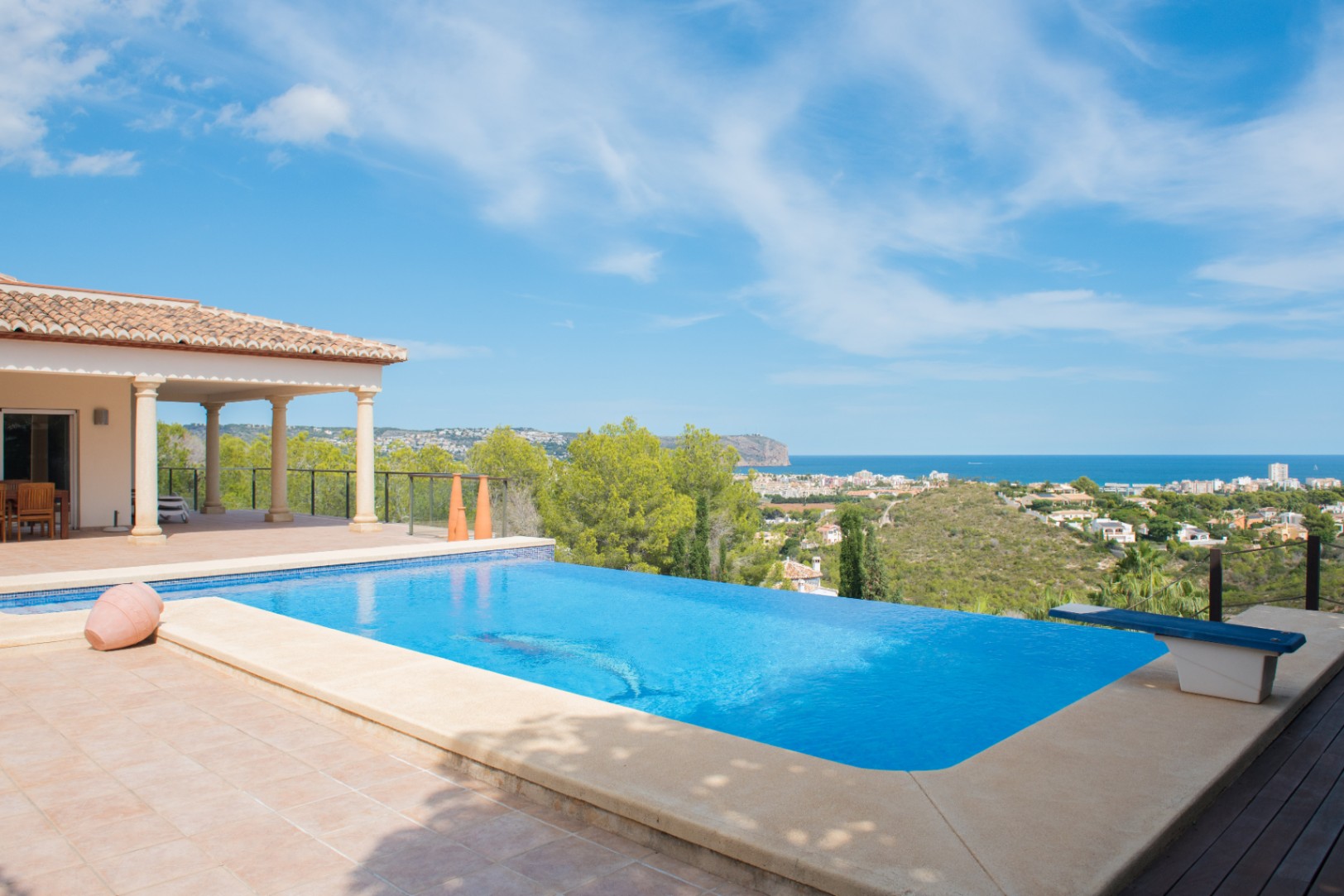 Piscina infinita con terraza de gres y trampolín, ofreciendo vistas panorámicas de la costa mediterránea y el paisaje boscoso circundante.