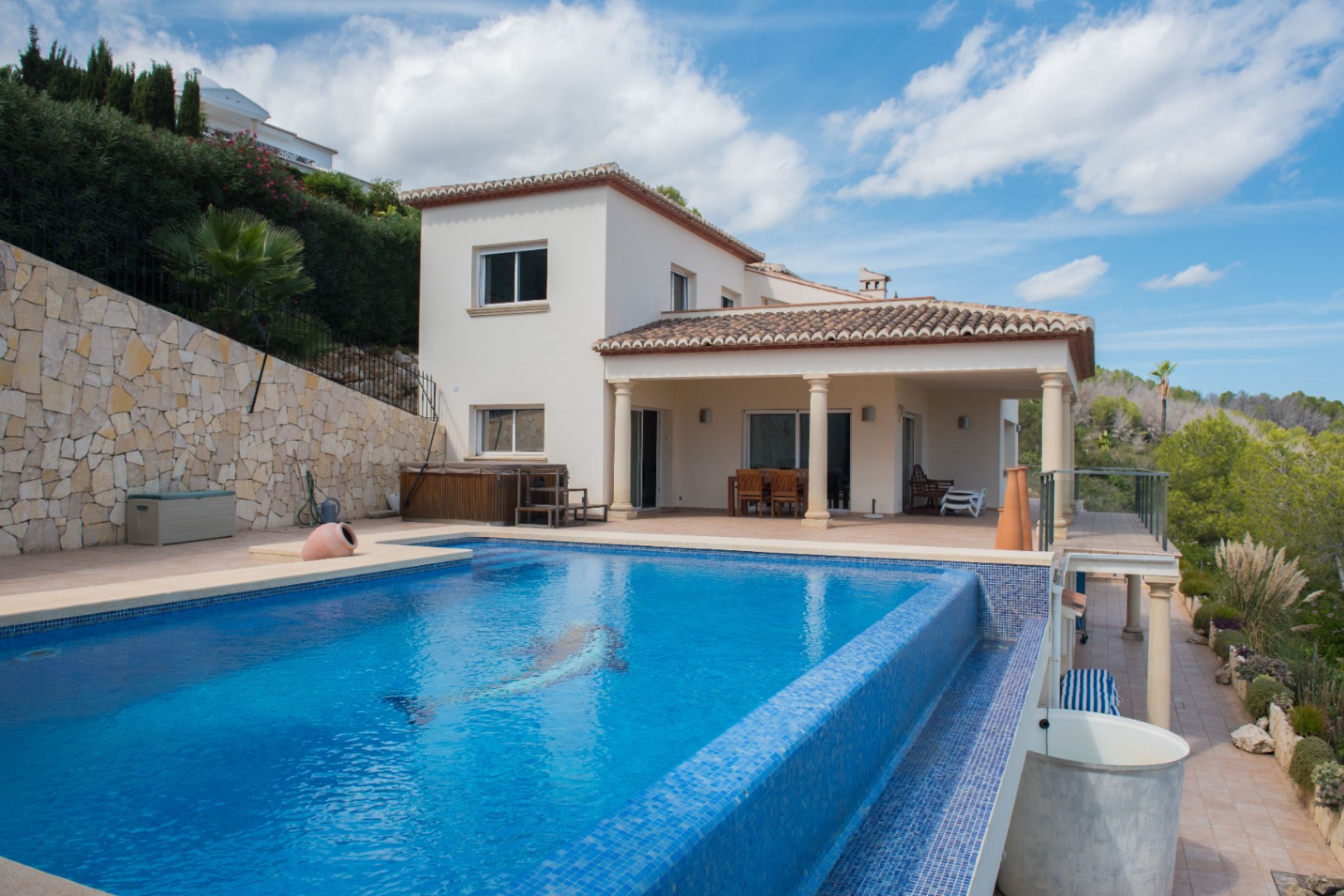 Vista exterior de una villa blanca con piscina de mosaico azul y motivo de delfín, junto a un porche cubierto con columnas y muros de piedra natural.