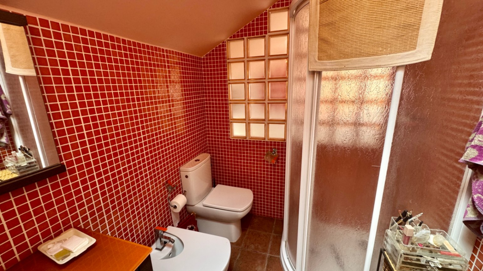 Baño con paredes de mosaico rojo, techo abuhardillado y ducha de esquina curva. Incluye sanitario, bidé y una ventana de bloques de vidrio que aporta luz natural.