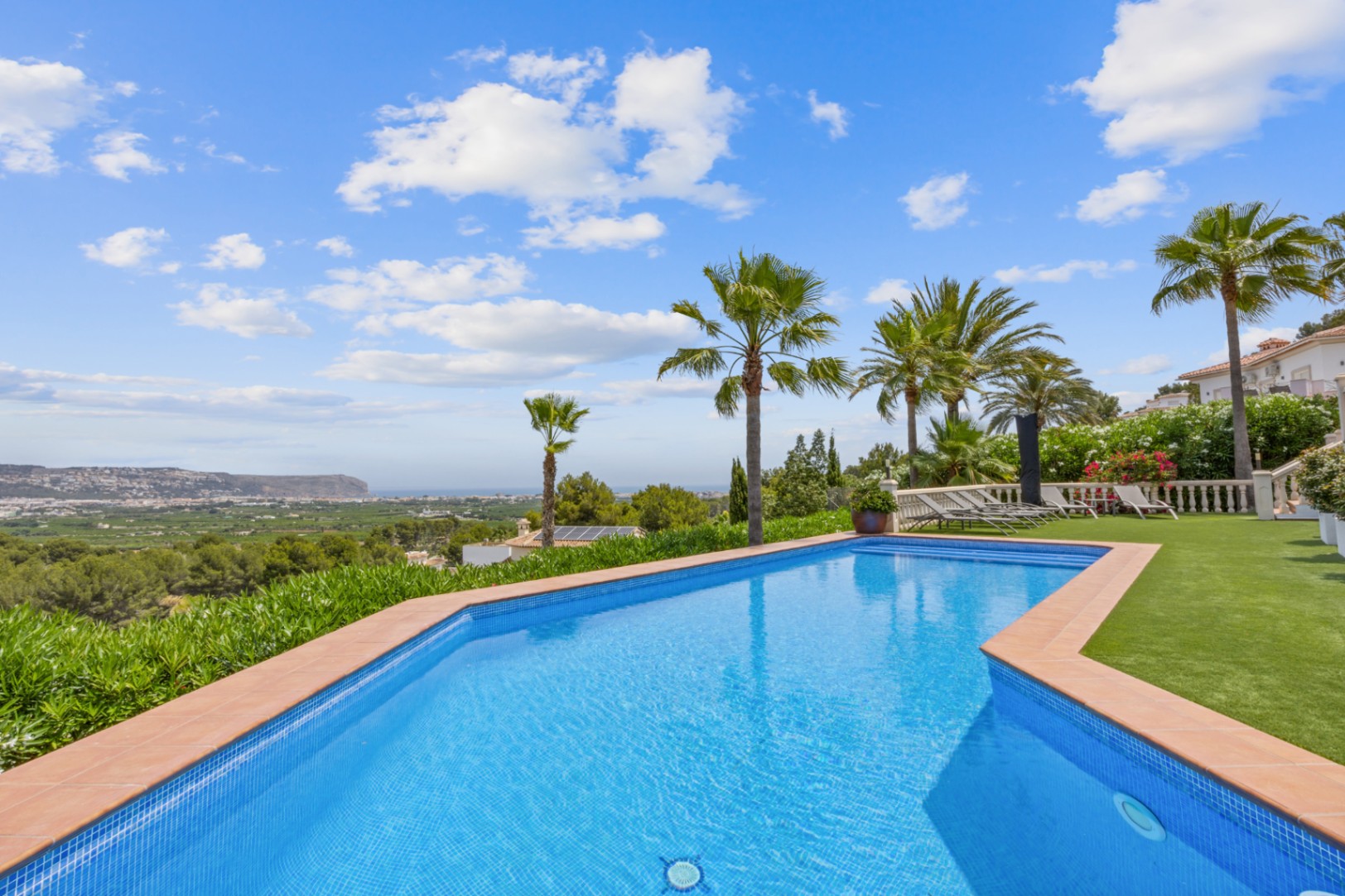 Piscina exterior con gresite azul y borde de terracota, con vistas panorámicas al valle y al mar, rodeada de palmeras y zona de césped artificial.
