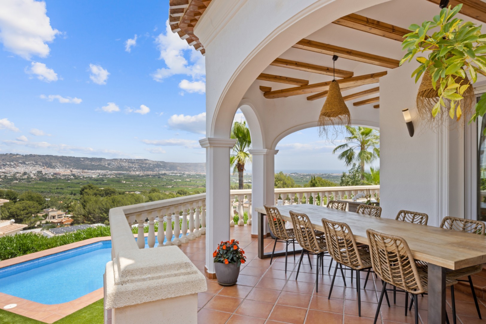 Terraza cubierta con vigas de madera, mesa de comedor exterior y vistas panorámicas al valle y al mar sobre la piscina privada.